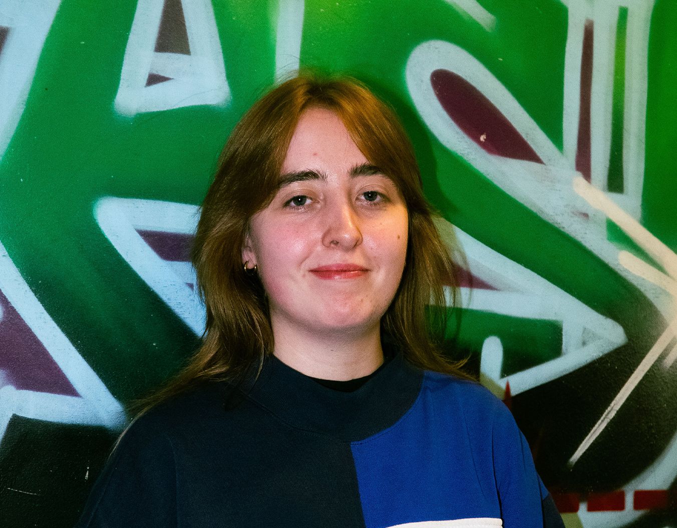 young adult female stood in front of a graffiti wall smiling at the camera