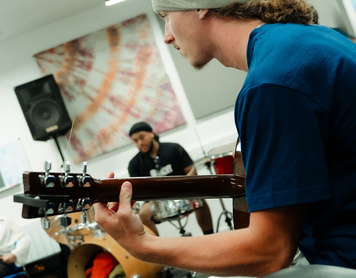 close up shot of a person playing guitar while another person is in the background sat on a chair
